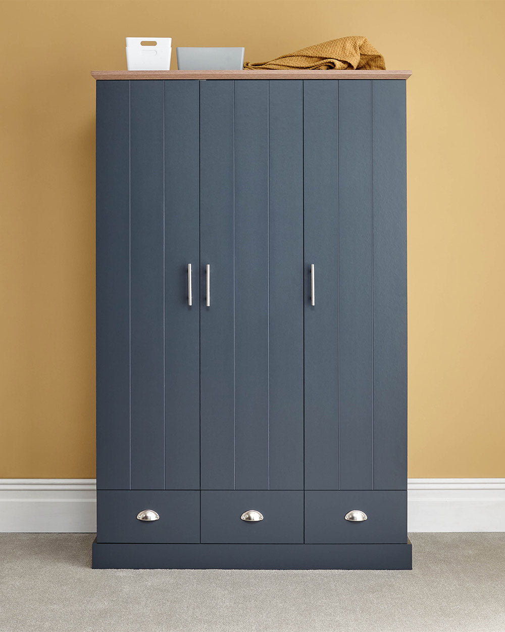 A 3 door wardrobe in modern slate blue offers ample storage space and elegant brushed steel handles. The wardrobe opens to reveal hanging space and a convenient shelf. It also features three drawers with cup handles. The bold slate blue colour complements any interior style and contrasts with the oak-effect top. The wardrobe is aesthetically pleasing and sturdy, with a deep base plinth. It comes flat packed for self-assembly. Delivery is available only to mainland UK, excluding certain postcodes.