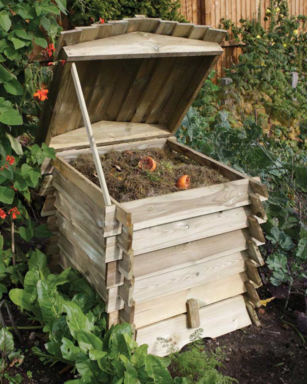 Garden compost bin in the shape of a beehive pictured in someone's garden. Rowlinson beehive composter with the lid up on the hinge with grass cuttings and orange peel inside