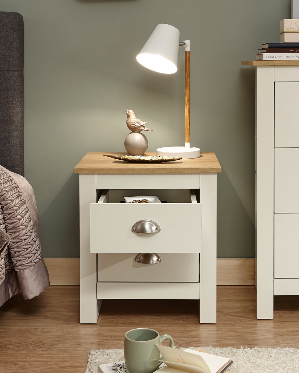 Lancaster bedside cabinet in cream with an oak effect top in a bedroom lifestyle setting with a lamp and decorative accessories on top with the top drawer open